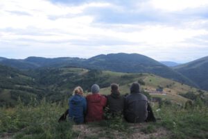 Vier Pfadfinder sitzen mit dem Rücken zur Kamera unter einer Zeltplane. SIe blicken über eine schöne Berg-Landschaft.