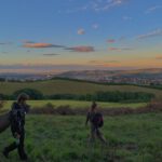 Zwei Menschen laufen auf einer Wiese entlang. Im Hintergrund erstreckt sich ein großes, hügeliges Tal hinter dem die SOnne untergeht und die Wolken Rosa färbt.