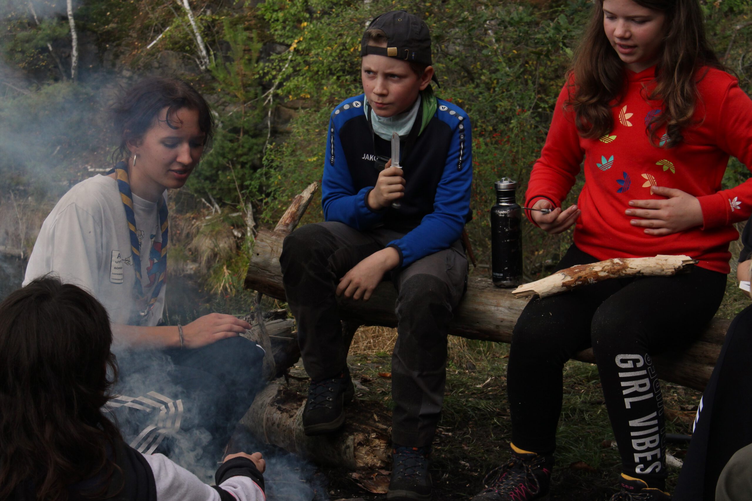 Zwei Kinder und eine Gruppenleitung sitzen auf Stämmen am Lagerfeuer, sie schnitzen. Der Qualm zieht durchs Bild.