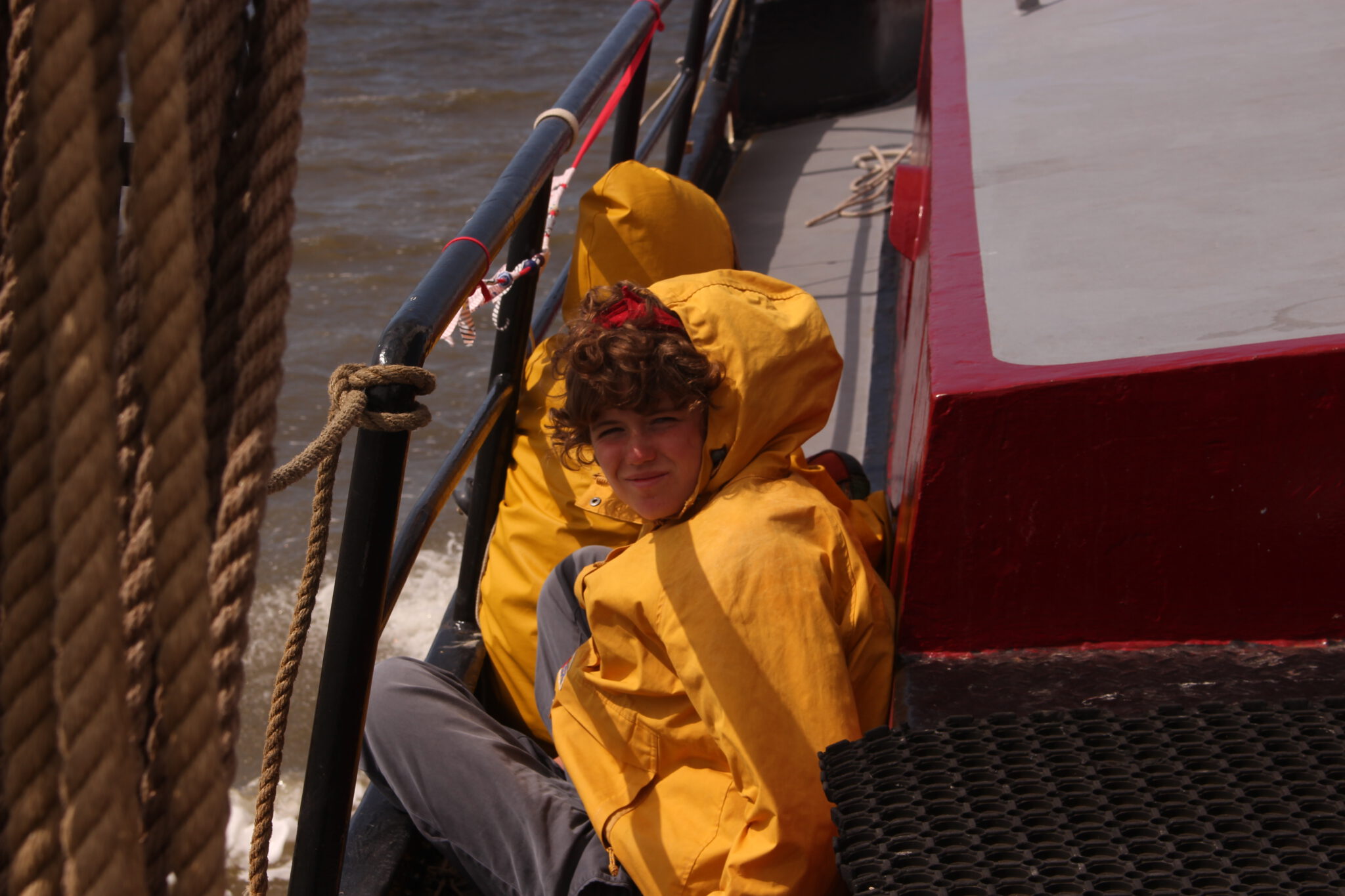 Eine Pfadfinderin in einer gelben Regenjacke sitzt im Gangway von einem Segelschiff und schaut in die Kamera.