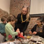 Zwei kleine Kinder und zwei Gruppenleitungen sind zu sehen. Die eine Leitung hilft einem Kind beim basteln. Im vordergrund sieht man viel Bastelmaterial auf einem Tisch ausgebreitet.